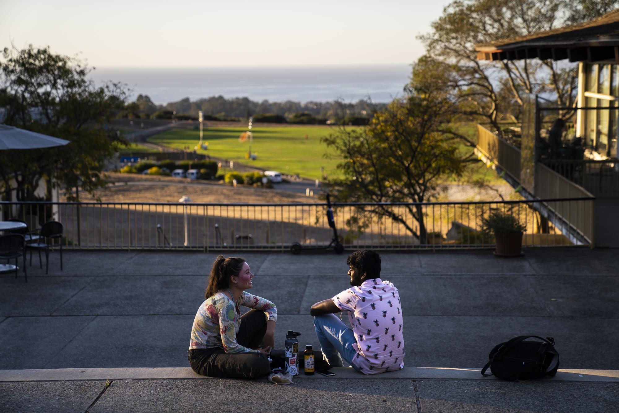 Two people sitting on the steps at Cowell College with a view of the athletics and recreation area in the background.